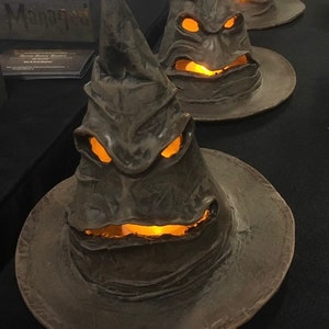 May include: Three gray, sculpted wizard hats with glowing orange light emanating from the eyes and mouth. The hats have a wide brim and a pointed top. The hats are displayed on a black surface.