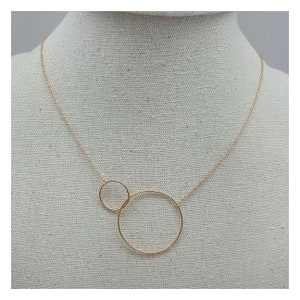 Può includere: Una collana d'oro con due cerchi intrecciati. Il cerchio più grande è in basso e quello più piccolo in alto. La collana è su uno sfondo di tessuto bianco.