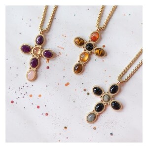 Peut inclure: Trois colliers croix dorés avec des pierres précieuses colorées. Une croix présente des pierres violettes et roses, une autre des pierres orange, noires et jaunes, et la troisième des pierres noires et grises. Chaque collier a une chaîne délicate.