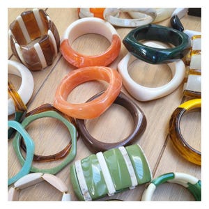May include: A collection of colorful, chunky bracelets in various shapes and designs. The bracelets feature shades of orange, brown, green, white, and yellow. Some have geometric shapes, while others are round or square. The bracelets are made of resin or plastic.