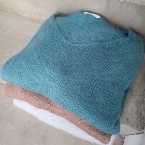 Peut inclure: Trois pulls pliés, de couleurs teal, beige et blanc. Le pull supérieur est teal avec un col en V. L'image met en valeur la texture douce des tricots. Les pulls sont empilés proprement, vus d'un angle légèrement surélevé.