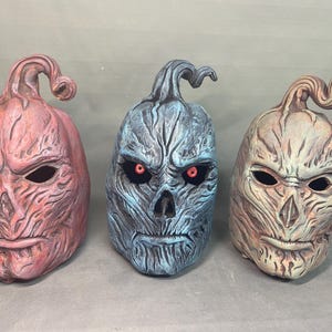 May include: Three Halloween masks with a carved pumpkin design. The masks are in shades of red, blue, and tan, each with a unique textured finish. The blue mask has glowing red eyes. The masks have a twisted stem on top.