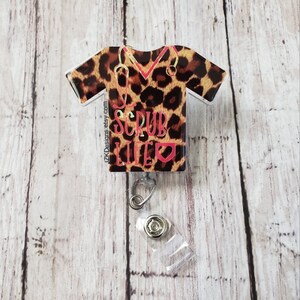 Peut inclure: Un badge rétractable en plastique transparent avec un haut de blouse imprimé léopard avec un stéthoscope et le texte "Scrub Life" en rose avec un cœur rose.