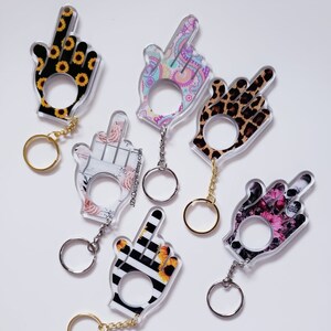 May include: A collection of acrylic hand-shaped keychains with various designs. The keychains feature patterns like sunflowers, leopard print, floral, and paisley. Each keychain has a gold or silver chain and ring for attaching keys.