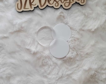 May include: Three circular objects, two white and one clear, are arranged on a fluffy white surface. Above them is a wooden sign with the text "JZK Designs" in a brown, handwritten-style font.