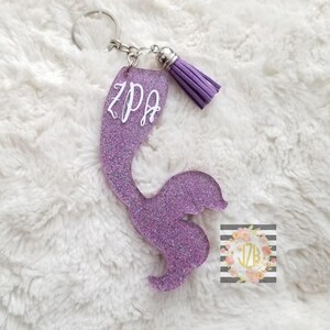 Peut inclure: Porte-clés en forme de queue de sirène en paillettes violettes avec un anneau de clé argenté et un pompon violet. La queue porte les lettres "ZPA" inscrites dessus.