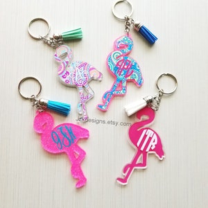 May include: Four pink flamingo keychains with different patterns and monograms. Each keychain has a silver key ring and a tassel.