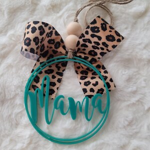 Peut inclure: Cercle turquoise avec le mot "Mama" d&eacute;coup&eacute; dans une police d'&eacute;criture cursive. Un n&oelig;ud en imprim&eacute; l&eacute;opard brun et beige est attach&eacute; au sommet du cercle avec deux perles en bois.