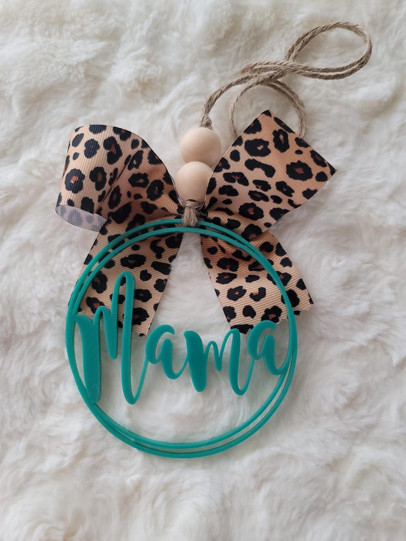 Peut inclure: Cercle turquoise avec le mot "Mama" d&eacute;coup&eacute; dans une police d'&eacute;criture cursive. Un n&oelig;ud en imprim&eacute; l&eacute;opard brun et beige est attach&eacute; au sommet du cercle avec deux perles en bois.