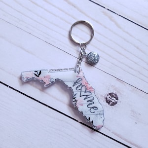 May include: A white and pink floral patterned Florida state keychain with a silver glitter charm and a silver key ring. The keychain says "Home" in silver glitter lettering.