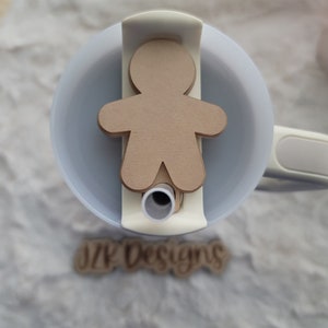 May include: A brown gingerbread man cutout is placed on a white lid of a travel mug. The mug has a white handle and a straw hole. The text "DIY Designs" is visible on the bottom of the image.