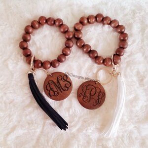 Peut inclure: Deux bracelets en perles de bois avec des porte-clés. Chaque porte-clés a un charme rond en bois avec des initiales gravées et un pompon. Un pompon est noir et l'autre est blanc.