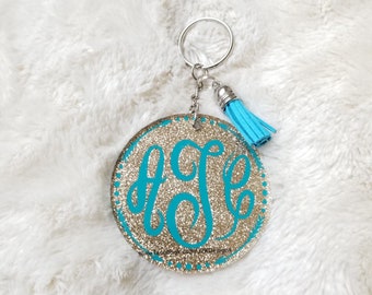 May include: A gold glitter circle keychain with a turquoise monogram and a turquoise tassel. The keychain has a silver ring and chain.