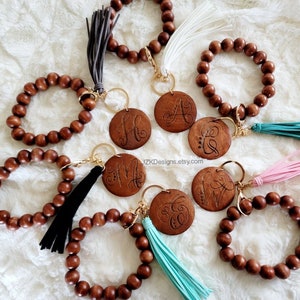 May include: Six wooden beaded bracelets with a keychain attached. Each bracelet has a different colored tassel and a wooden charm with a letter engraved on it. The charms are engraved with the letters A, C, H, M, V, and W.