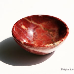 May include: A small, round bowl made of red and white marbled stone. The bowl has a smooth, polished surface and a natural, organic appearance. The bowl is empty and sits on a white surface.