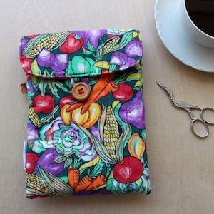 May include: A rectangular quilted fabric pouch featuring a vibrant vegetable print with corn, carrots, peppers, and leafy greens on a dark green background. The pouch has a fold-over flap secured by a single round wooden button.
