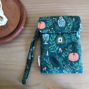 May include: A quilted fabric pouch with a wrist strap featuring a dark green background printed with orange pumpkins, white spiderwebs, black crows, candles, and glass jars containing plants and insects. A small white fabric tag is on the side.