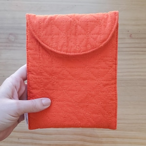 May include: An orange quilted fabric case with a flap closure. The case has a geometric diamond pattern created by the stitching. The case is held by a hand, and the background is a light wood surface.