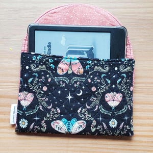 May include: A quilted fabric sleeve for an e-reader featuring a black background with a pattern of moths, stars, and flowers. The sleeve has a pink speckled interior flap and a small white tag labelled Stitched & Unbound on the side.