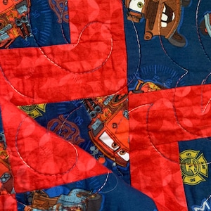 May include: A red, blue, and brown quilt with a Cars theme. The quilt features images of Lightning McQueen and Mater, along with the text "Race Day" and "50".