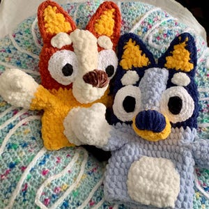 May include: Two handmade crochet dog puppets. One is orange, yellow, and white, the other is blue, yellow, and white. Both have large black eyes and are laying on a light blue and white patterned blanket. The puppets are soft and plush.