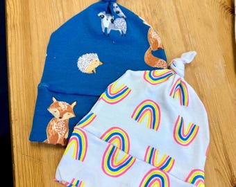 May include: Two baby knot hats. One is blue with a fox, hedgehog, and sheep print. The other is white with a rainbow pattern in pink, yellow, and blue. The hats are on a wooden surface.