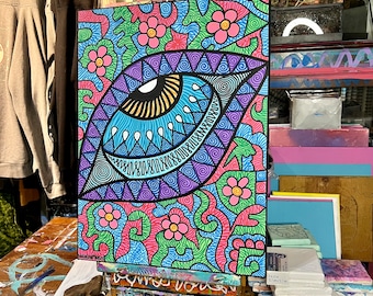 May include: A colorful abstract painting on canvas with a large eye design in the center. The eye is surrounded by pink flowers and swirls of blue, green, and purple. The painting is on an easel in an art studio.