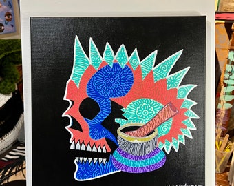 May include: A black canvas painting with a colorful skull design. The skull is outlined in white and filled with blue, red, and green patterns. The skull is wearing a crown of spikes and has a small bowl with a spoon on top of its head. The painting is signed "WILDFIREMOSES 2023" in the bottom right corner.