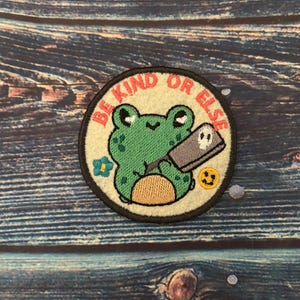 May include: A green frog holding a knife with a skull on it. The frog is surrounded by a white circle with the text "Be Kind Or Else" in pink.