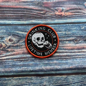 Peut inclure: Un écusson rond avec un fond noir et une bordure orange. L'écusson représente un crâne blanc buvant dans une chope. Le texte "DRINKING CLUB LIFETIME MEMBER" est en blanc autour du crâne.
