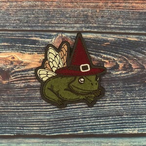 Puede incluir: Parche bordado con una rana verde que lleva un sombrero de bruja burdeos con una hebilla blanca. La rana tiene alas de hada de color beige y blanco. El parche está contorneado en marrón oscuro sobre un fondo de madera.