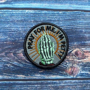 May include: Round embroidered patch with the text "PRAY FOR ME, I'M PETTY". The patch features a green skeleton hand in the center, surrounded by an orange sunburst design. The patch has a black border and is set against a blue wooden background.
