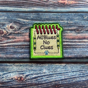 May include: Embroidered patch featuring a notepad design with the text "All Blues No Clues." The patch has a green border, red spiral binding, and a blue paw print. Two green and yellow pencils frame the notepad.