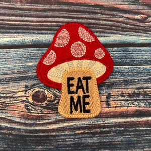 Peut inclure: Patch en forme de champignon rouge et blanc avec les mots "Eat Me" sur un fond beige.