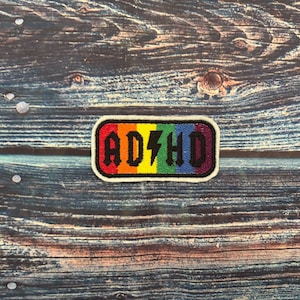 Puede incluir: Un parche rectangular con las letras "ADHD" en negro, con un rayo en el centro. Las letras están sobre un fondo de arcoíris, y el parche tiene un borde blanco. El parche está sobre una superficie de madera.