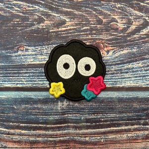 May include: A black, cloud-shaped patch with large white eyes and a smiling mouth. The patch is decorated with three colourful star-shaped accents in yellow, pink, and teal. The patch is likely for clothing or accessories.