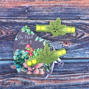 May include: A face mask with a green and gray floral pattern and the text "Die Mad About It". The mask has two green leaf-shaped attachments with yellow buttons.