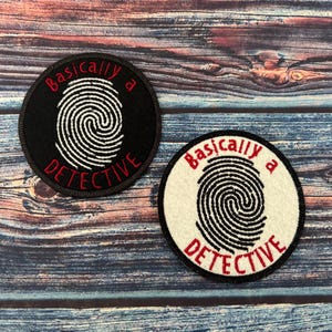 Op de afbeelding: Twee geborduurde patches in zwart en wit met de tekst "Basically a Detective" en een vingerafdrukdesign. De patches bevinden zich op een houten achtergrond.
