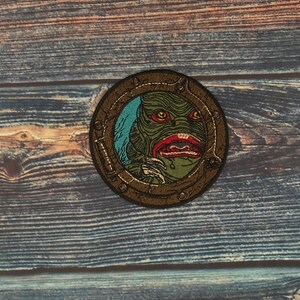 Può includere: Toppa rotonda raffigurante una creatura verde con labbra e occhi rossi, su sfondo blu all'interno di una cornice circolare marrone. La toppa ha un'estetica vintage e retrò, adatta a giacche o borse.