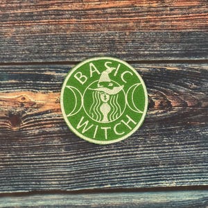 Peut inclure: Un écusson vert circulaire avec les mots "BASIC WITCH" en lettres blanches. Le motif comprend une sorcière portant un chapeau et des croissants de lune. L'écusson est sur une surface en bois.