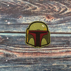 May include: Embroidered patch depicting a bounty hunter's helmet. The helmet is predominantly olive green with a red and black visor. Gold and yellow details are present on the side and top. The patch is set against a rustic, wood-grain background.