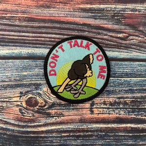 May include: A round, blue and black embroidered patch with a pink outline. The patch features an ostrich with its head tucked under its wing and the text "Don't Talk To Me".