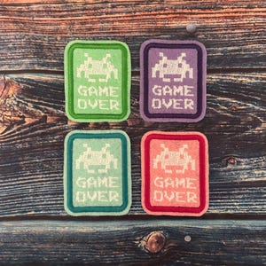 May include: Four felt patches featuring a pixelated alien and the text "GAME OVER". The patches are in green, purple, teal, and pink.