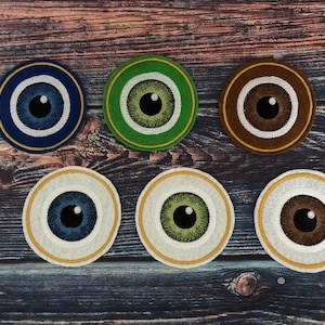 Puede incluir: Seis parches bordados con forma de ojo en azul, verde y marrón. Cada parche presenta un diseño detallado del iris y la pupila, rodeado por un borde blanco y dorado. Los parches están dispuestos sobre una superficie de madera oscura.