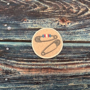 May include: A round, tan patch featuring two safety pin illustrations. One pin has a decorative bead design with red, white, and blue beads. The patch is set against a rustic, blue and brown wooden background.