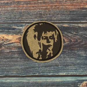 May include: A circular patch with a portrait design in black and tan. The portrait is enclosed within a black border. This patch is likely intended for attachment to garments or accessories.