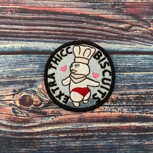 May include: A round, embroidered patch with a white cartoon character wearing a chef's hat and red shorts. The text "EXTRA THICC BISCUITS" is embroidered around the character.
