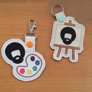 Könnte beinhalten: Zwei weiße Schlüsselanhänger mit einem Cartoon-Künstler-Thema. Der eine Schlüsselanhänger hat die Form einer Farbpalette mit bunten Farbspritzern. Der andere Schlüsselanhänger hat die Form einer Staffelei mit einem Gemälde. Beide Schlüsselanhänger zeigen eine schwarze Afro-Frisur und einen Bart.
