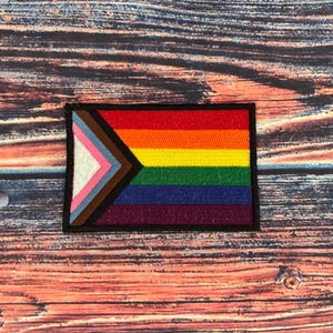 May include: Rectangular patch featuring the Progress Pride flag design. The flag includes the traditional rainbow stripes, along with a chevron of colors representing transgender, marginalized, and people of color communities. The patch has a black border.