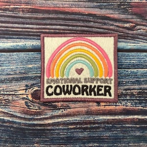 May include: A white felt patch with a rainbow design and the text "Emotional Support Coworker". The rainbow is made of pink, orange, yellow, green, blue, and purple thread. A small red heart is in the centre of the rainbow.